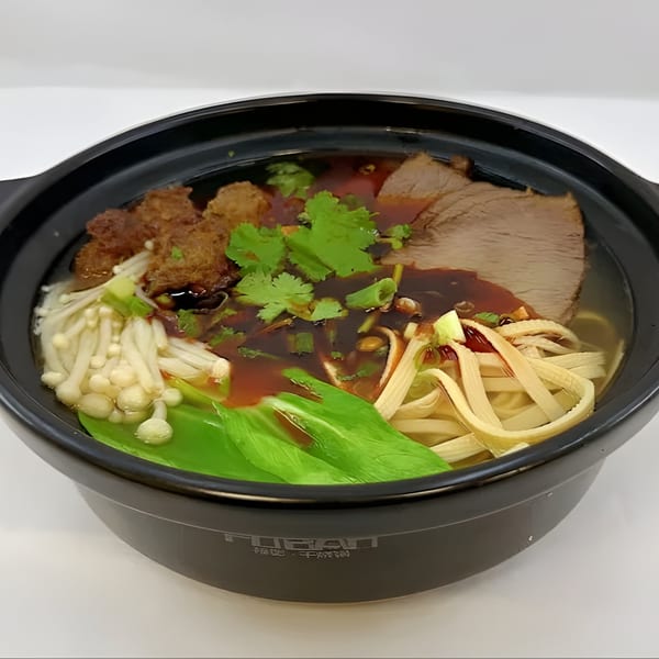 A black bowl of Turkish noodle soup with beef, mushrooms, greens, and a dark sauce.