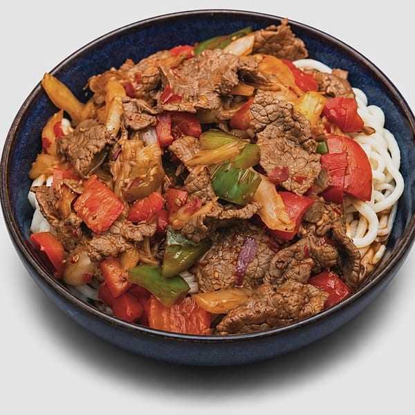 A blue bowl filled with stir-fried beef, red and green bell peppers, onions, and thick white noodles.